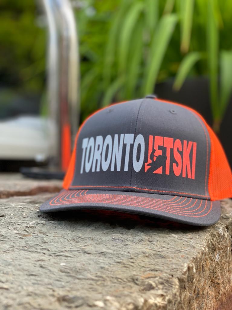 Toronto Jet Ski Trucker Hat: Front panel in grey with white embroidered 'Toronto' and orange 'jetski' logo. Back features breathable orange mesh material. Orange stitching accents the brim, adding a coordinated touch to this stylish watersport accessory.